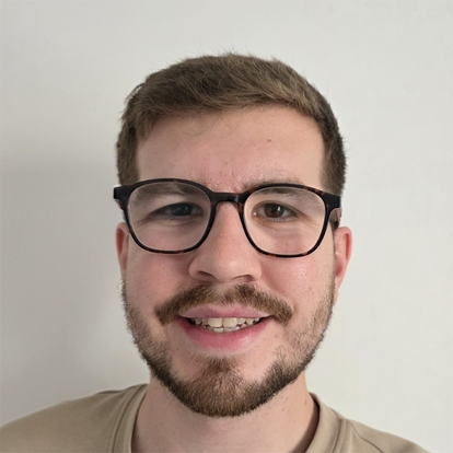 Photograph of Willis. He has short brown hair, glasses, and short facial hair. He is smiling while wearing a grey and T-shirt and looking into the distance.